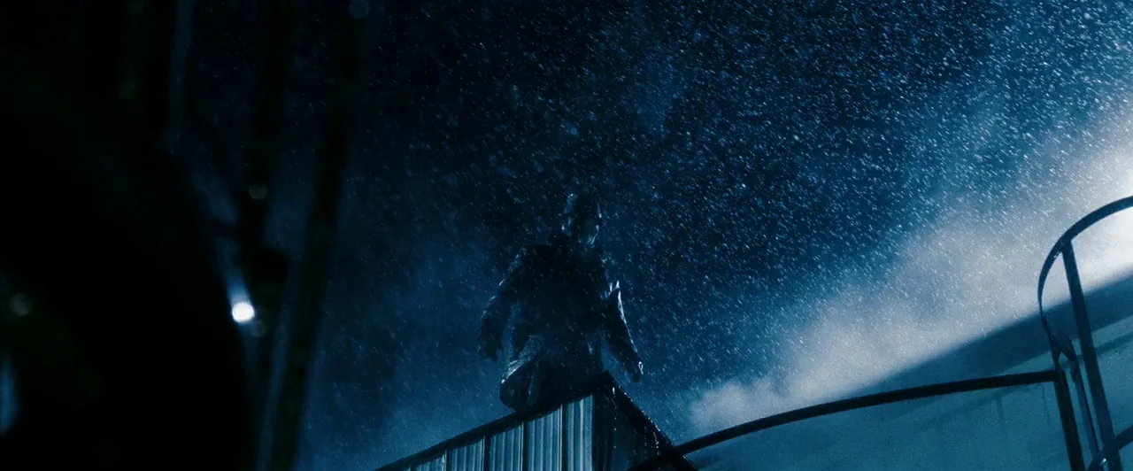 Watchmen (2009) movie still: wide — A dark rooftop structure is illuminated by a harsh light source during heavy rain. A solitary man in a dark coat…