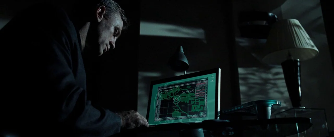 Casino Royale (2006) movie still: medium — A dark, dimly lit office at night contains a glass-topped desk and a table lamp. A man sits at the desk…