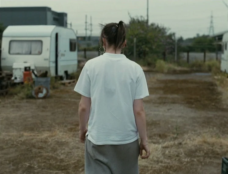 Fish Tank (2009) movie still: medium — A desolate, open patch of dirt and dry weeds sits between two white trailers. A person walks forward away…
