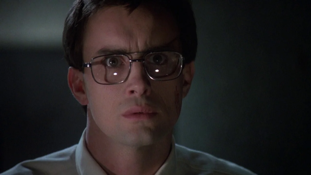 Re-Animator (1985) movie still: closeup — A dimly lit indoor space with dark, indistinct surroundings. A man with glasses stares forward with wide,…