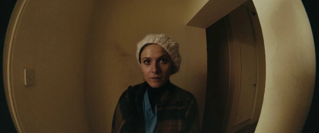Saint Maud (2019) movie still: medium — The interior of a doorway is viewed through a high-distortion fisheye security lens. A woman stands in the…