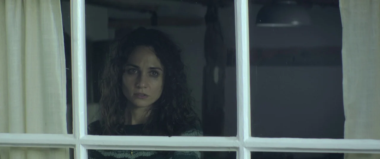 Lord of Misrule (2023) movie still: medium — A dark indoor space viewed through a white grid window frame at night. A woman with long curly hair…