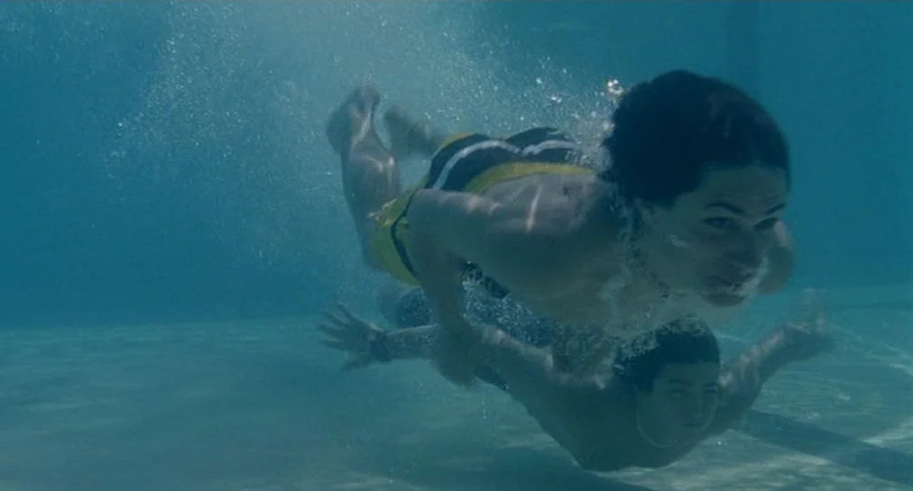 Y Tu Mamá También (2001) movie still: fullbody — Two people swim together underwater in a deep blue pool with bubbles forming around them. The…