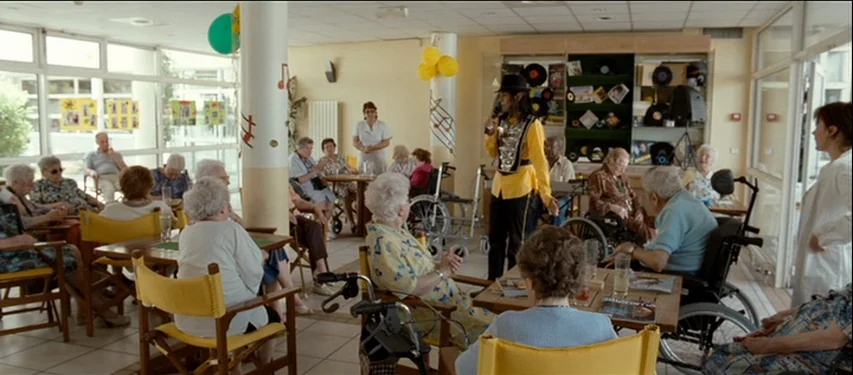 Mister Lonely (2007) movie still: wide — A nursing home day room features floor-to-ceiling windows, white support pillars, and elderly residents. A…
