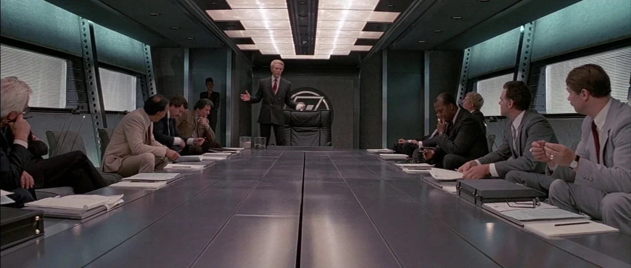 A View to a Kill (1985) movie still: wide — A modern 1980s boardroom features a long metallic table and recessed panel lighting. Multiple men in…