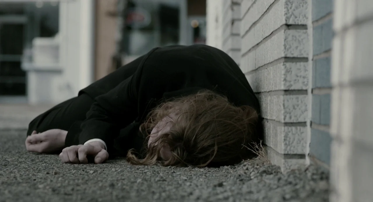 Antiviral (2012) movie still: medium — A person lies prone on a gravelly sidewalk next to a white brick wall. The subject is wearing a black suit…