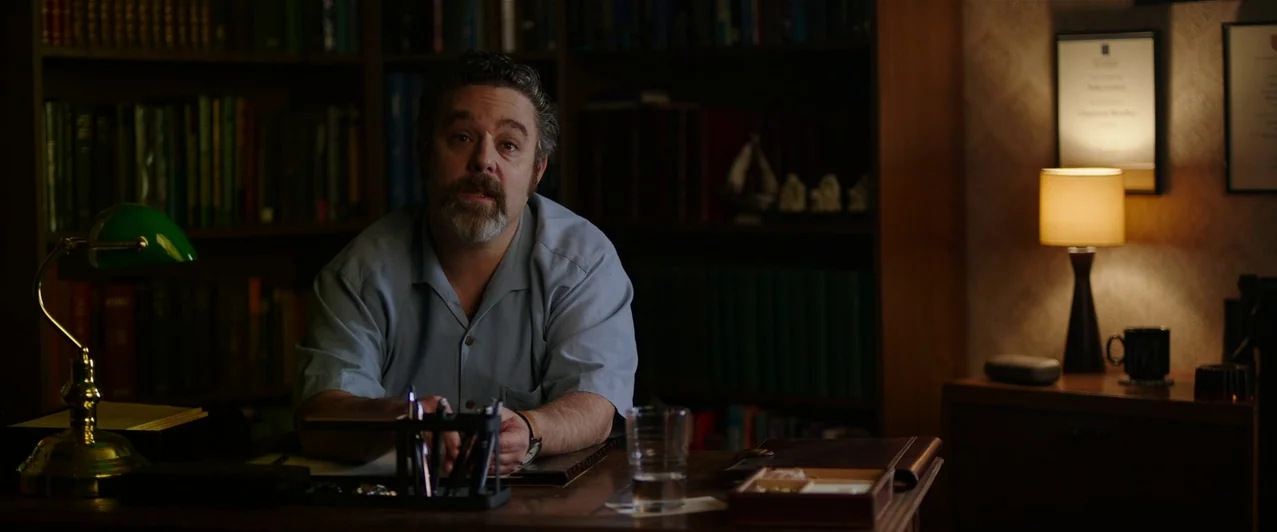 Ghost Stories (2017) movie still: medium — A wood-paneled room is lined with dark bookshelves filled with various hardbound books. A man with a…