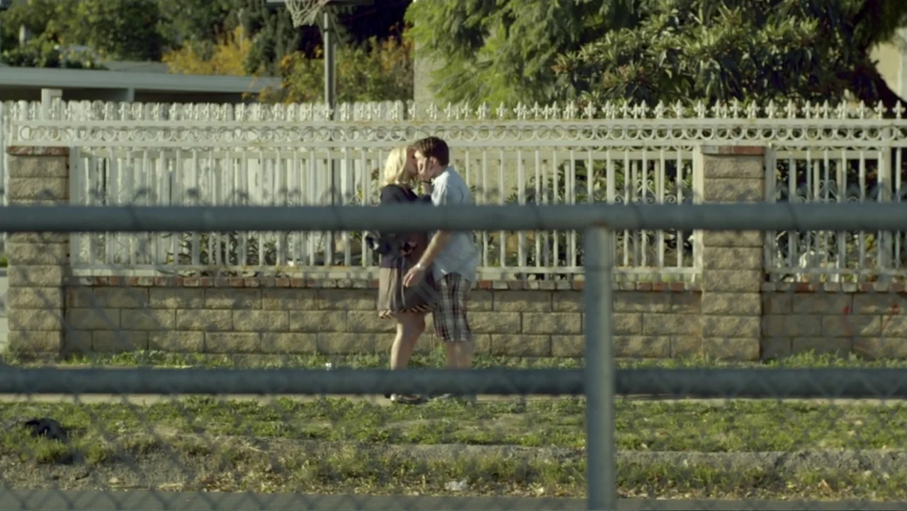 Free Samples (2013) movie still: fullbody — A residential sidewalk bordered by a white wrought iron fence and brick base. A man and woman in casual…