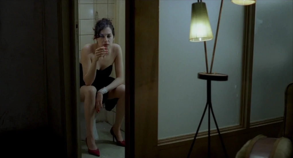 Anatomy of Hell (2004) movie still: medium — A woman sits in a tiled bathroom stall seen through an open doorway. She holds a cigarette in one hand…