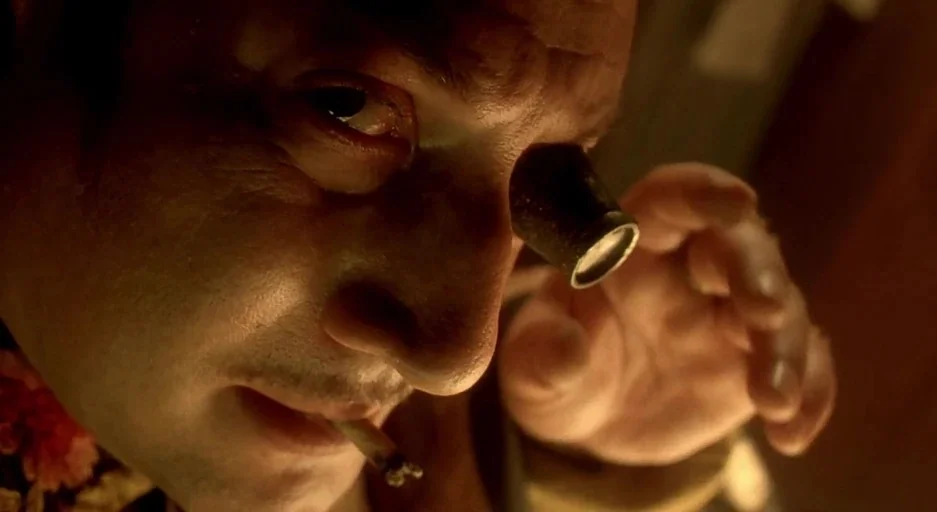 The City Of Lost Children movie still: closeup — A dimly lit, cramped workspace contains a man examining an object closely. He holds a jeweler's…