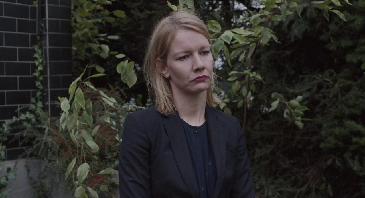 Toni Erdmann (2016) movie still: medium — An outdoor space features a dark tiled wall and dense foliage in the background. A woman in a black blazer…