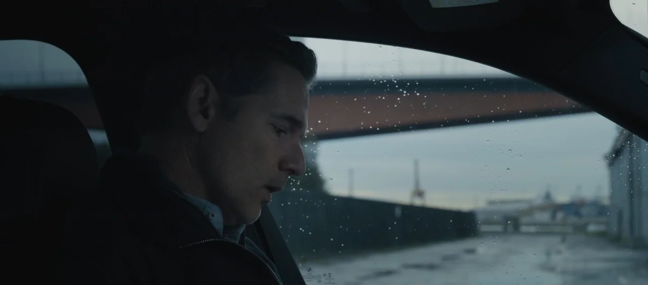 Force of Nature: The Dry 2 (2024) movie still: medium — A man sits inside a vehicle while parked near an industrial waterfront with a bridge. The…