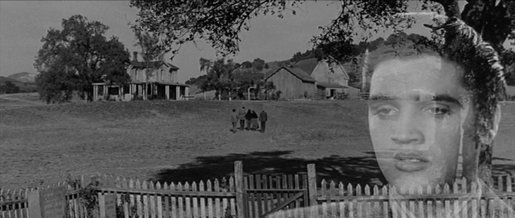 Love Me Tender movie still: wide — A rural landscape features a farmhouse and barns near a fenced graveyard. A large, transparent portrait of a man…
