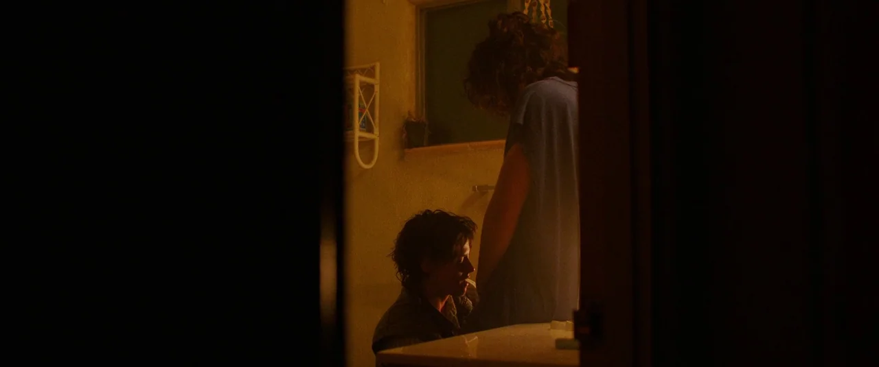 Love Lies Bleeding (2024) movie still: medium — A dimly lit room with walls painted a mustard yellow and a small window frame. A person in a blue…