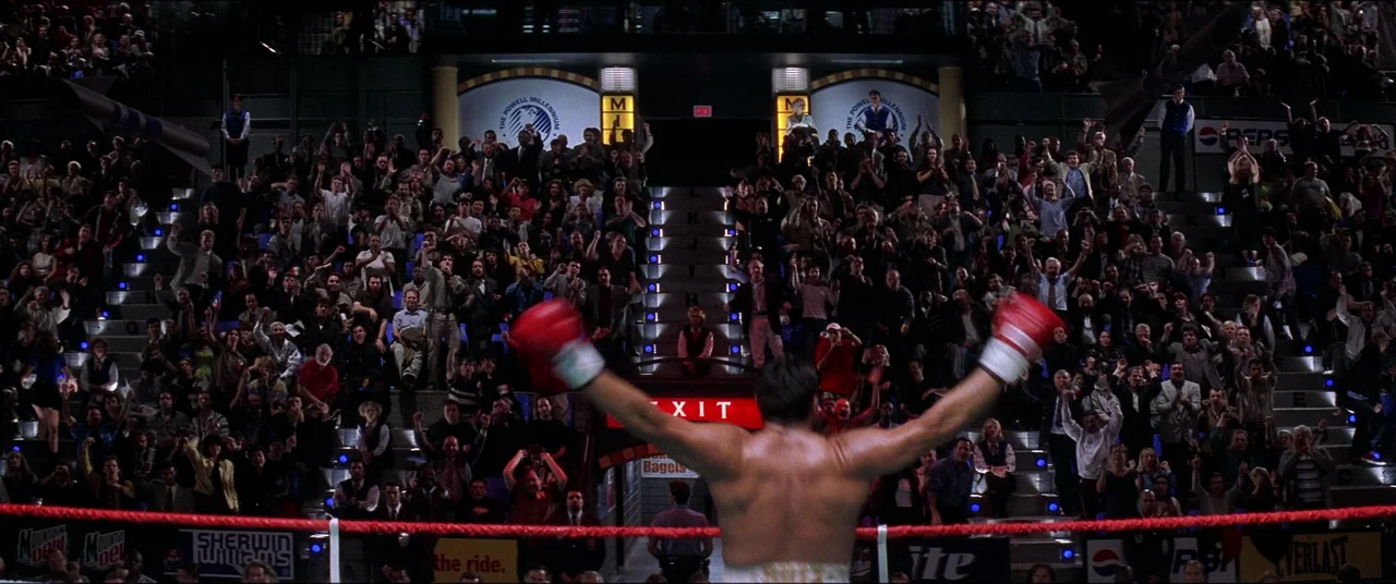 Snake Eyes (1998) movie still: fullbody — A crowded indoor sports arena viewed from behind a boxer in the ring. A large audience fills the tiered…