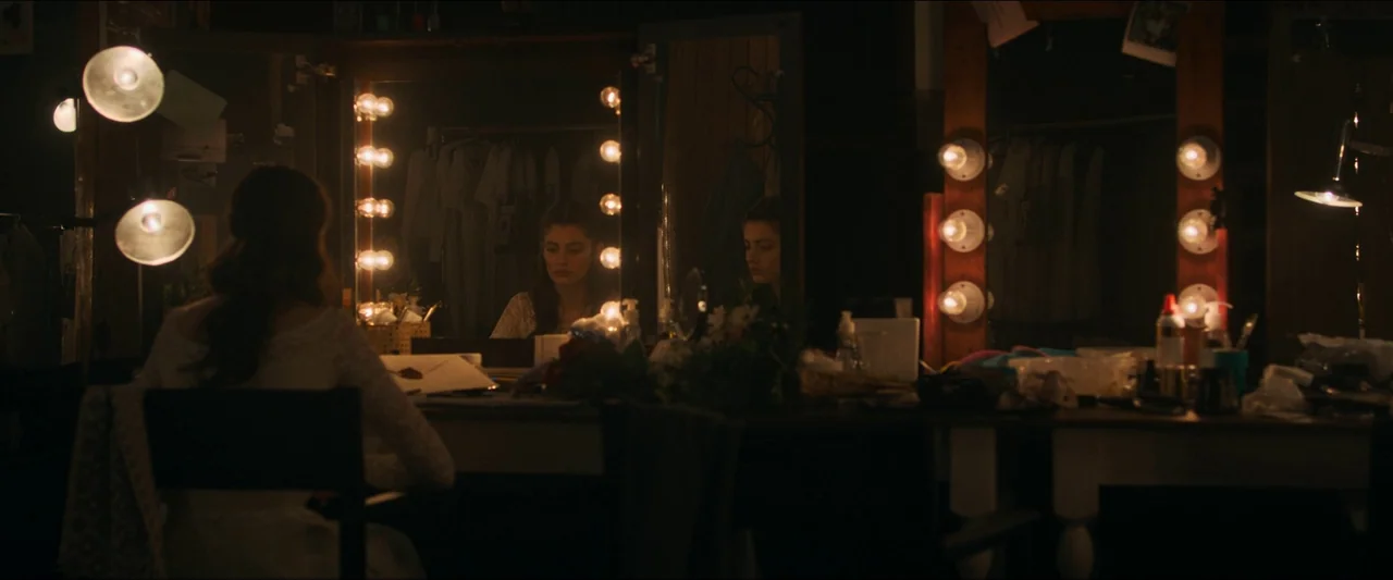 Birds of Paradise (2021) movie still: medium — A dimly lit vanity area with mirrors surrounded by warm lightbulbs and personal clutter. A woman in a…