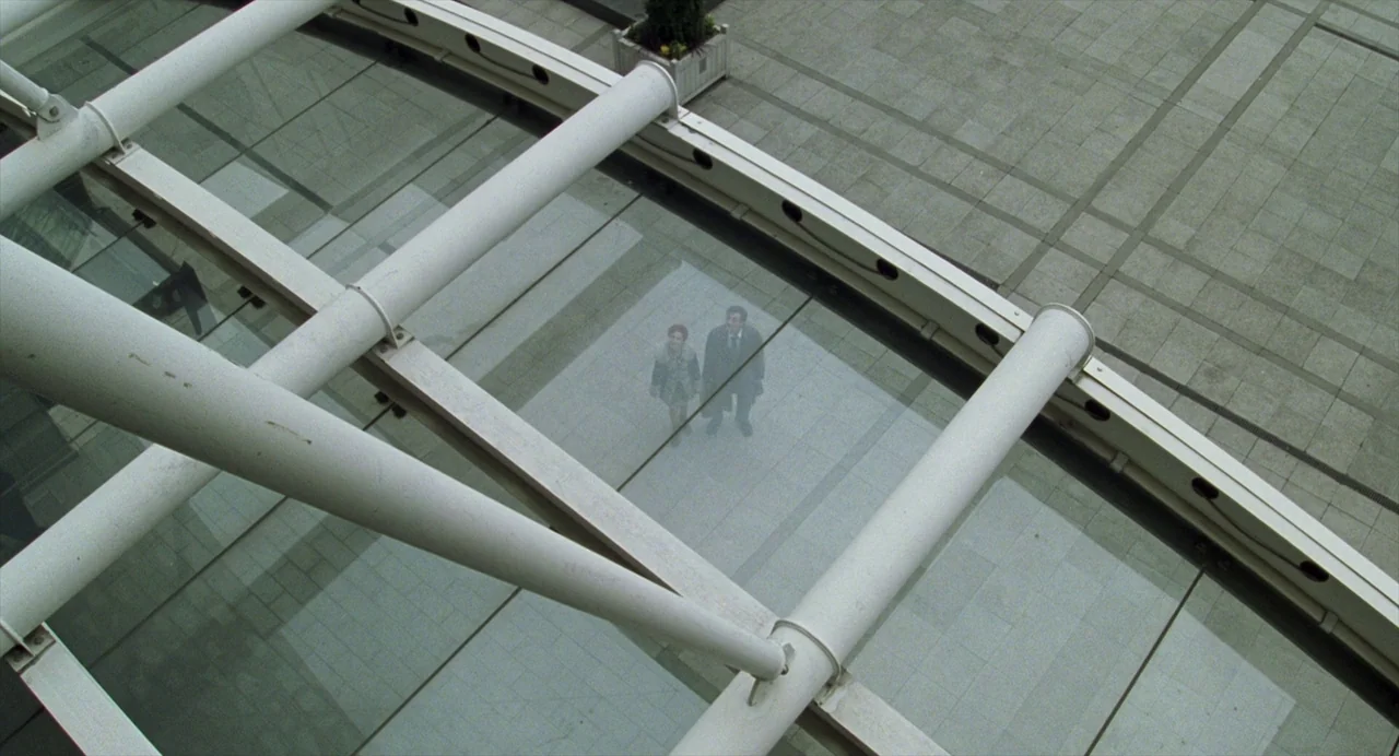 Closer (2004) movie still: wide — A vast modern paved plaza features large white structural pipes extending across the frame. Two individuals stand…