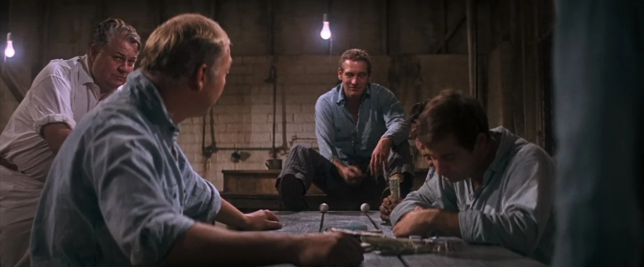 Cool Hand Luke (1967) movie still: medium — A dimly lit room with bare brick walls and hanging light bulbs. Several men sit around a wooden table…