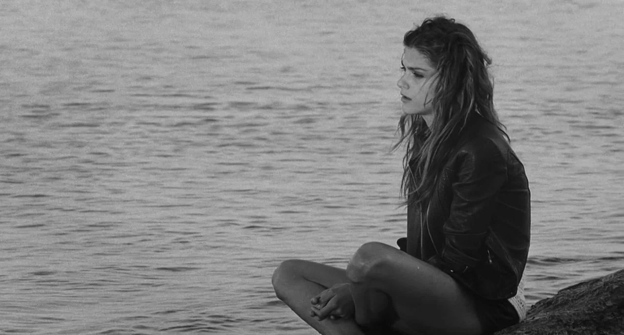The Legend Of Kaspar Hauser (2012) movie still: medium — A woman sits at the edge of a calm body of water. The shoreline is visible in the…