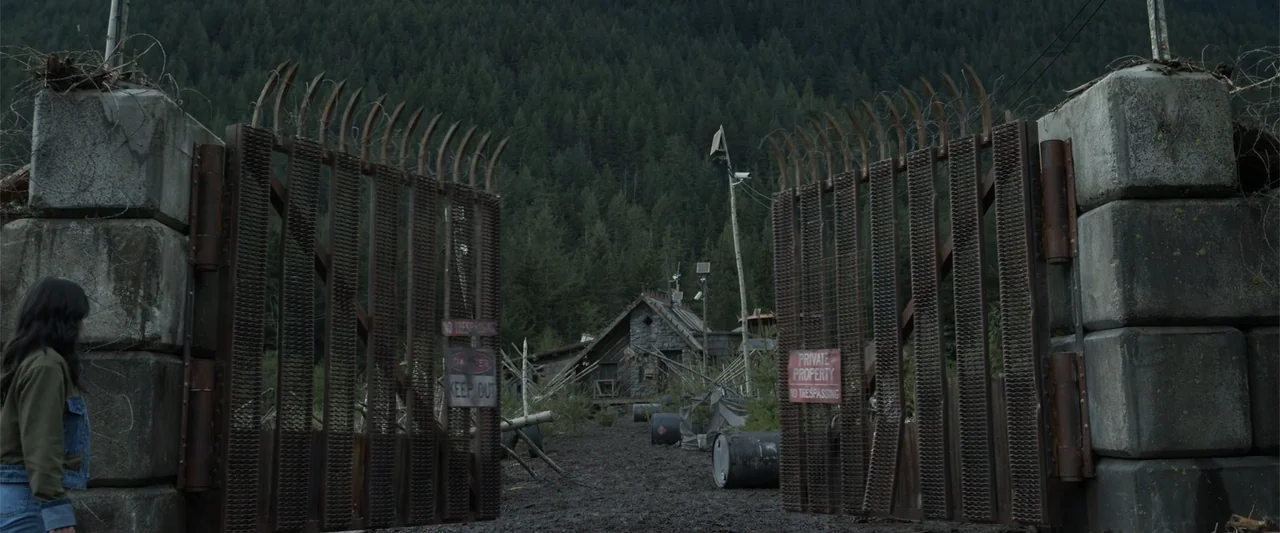 Final Destination Bloodlines (2025) movie still: medium — An open, rusty metal gate with barbed wire stands before a forested mountain path. A woman…