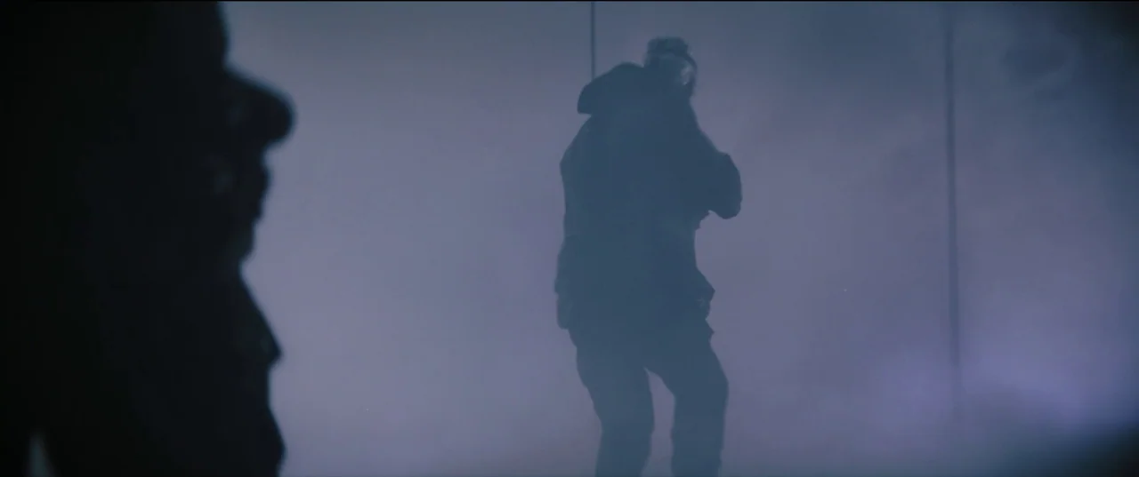 The Batman (2022) movie still: medium — An expanse of hazy, greyish blue space is obscured by thick atmospheric fog. A silhouetted figure stands in…