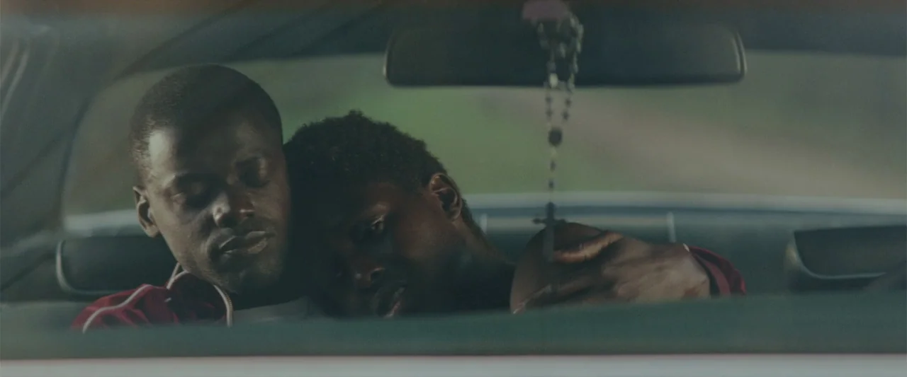 Queen & Slim (2019) movie still: medium — Two men sit together in the front seats of a moving car during daylight. One man leans his head onto the…