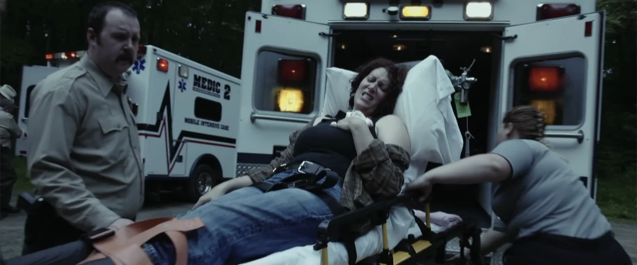 Darkness Waits (2020) movie still: medium — An emergency medical scene outdoors at the back of a parked white ambulance. A woman lies on a gurney…