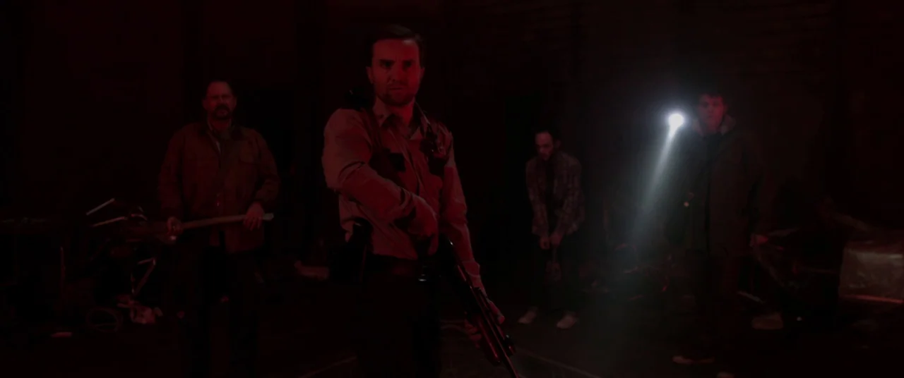 The Void (2016) movie still: medium — A dark subterranean space filled with clutter and indistinct shapes cast in red light. Three men stand near…