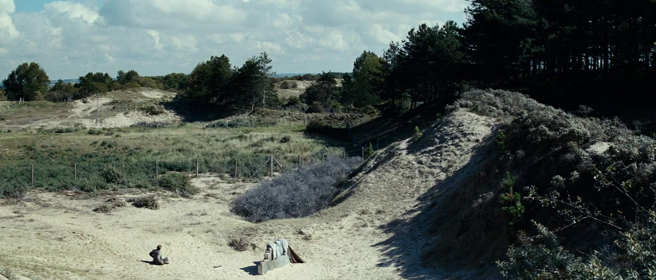 Outside Satan (2011) movie still: wide — A vast coastal dune landscape features sandy slopes, sparse shrubs, and dense evergreen trees. A lone…
