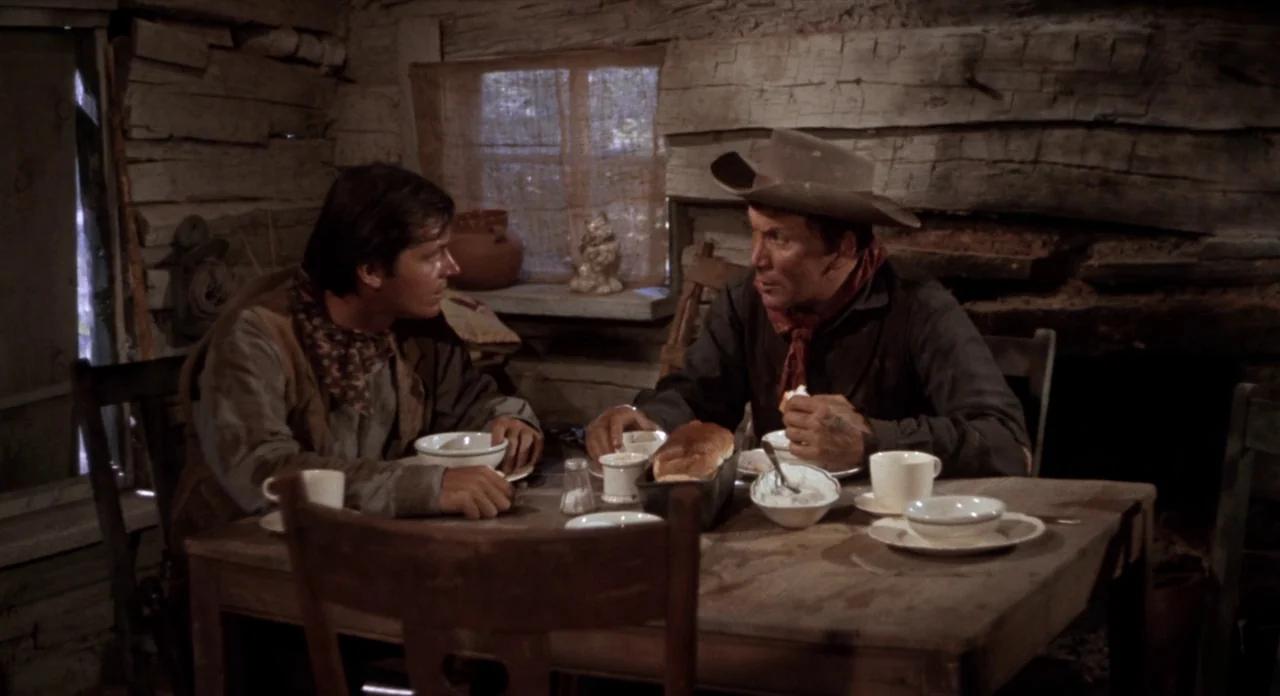 Ride In The Whirlwind (1966) movie still: medium — A rustic log cabin interior features a dining table made of weathered wood. Two men sit across…