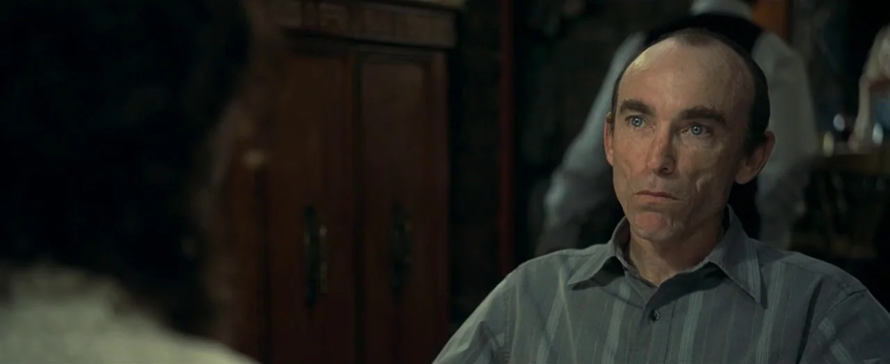 Little Children (2006) movie still: medium — A dimly lit interior space with dark wooden cabinets in the background. A thin, balding man sits and…