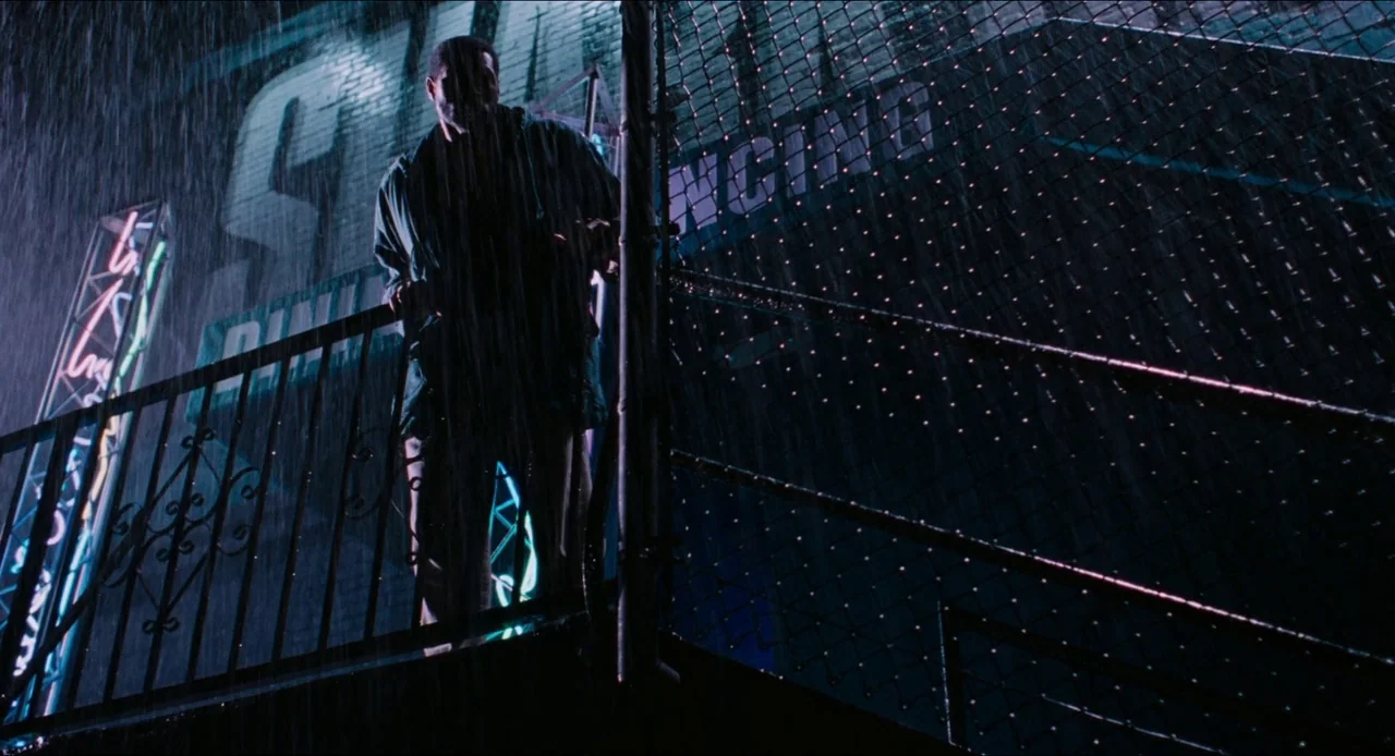 Deep Cover (1992) movie still: fullbody — A rain-slicked metal fire escape platform stands against a brick wall with neon signs. A man in a dark…