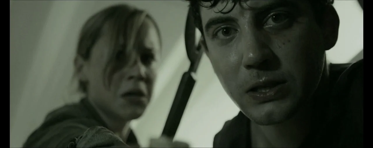 Turbulence (2025) movie still: closeup — A dimly lit industrial hallway features a man and woman looking down at camera. They are sweating and…