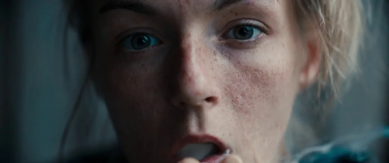 Silver Haze (2023) movie still: closeup — A close-up shot of a young woman's face in a dimly lit indoor space. She looks into the camera while…