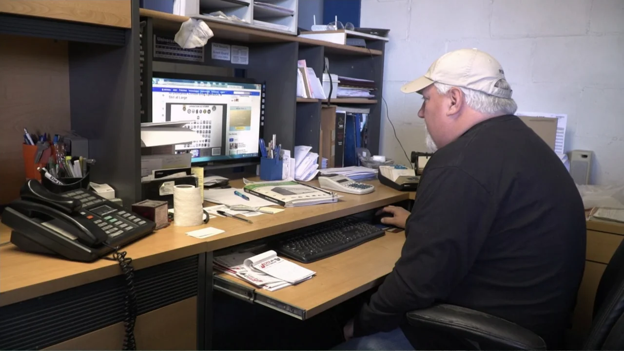 Meth Storm (2017) movie still: medium — A small office space features a wooden desk cluttered with paperwork and electronics. A man sits at his…