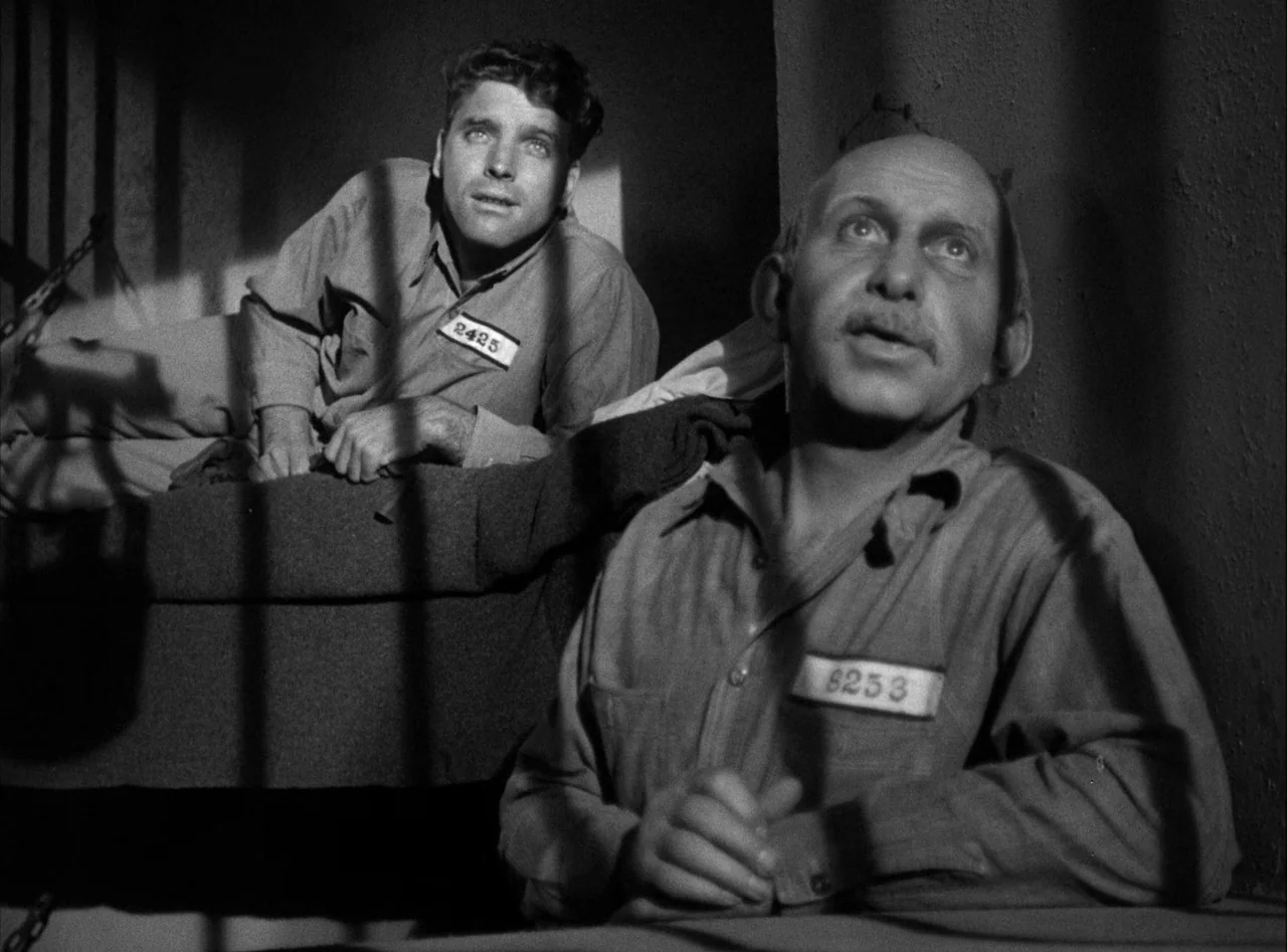 The Killers (1946) movie still: medium — A stone-walled room features barred windows that cast sharp shadows across two men. The individuals occupy…