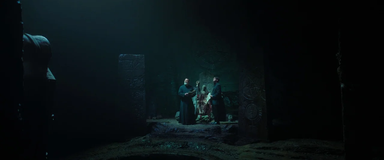 The Pope’s Exorcist (2023) movie still: wide — A dark, subterranean stone chamber features large carved pillars and a weathered central figure. Two…