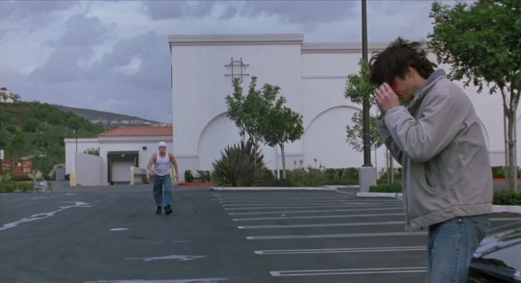 Brick (2005) movie still: wide — A sprawling empty asphalt lot sits before a large, stark white commercial building. One man runs across the…