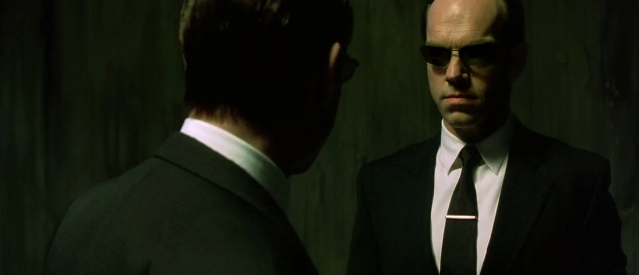 The Matrix Reloaded (2003) movie still: medium — A dark, nondescript room with textured walls provides a minimal backdrop for the figures. A man in…