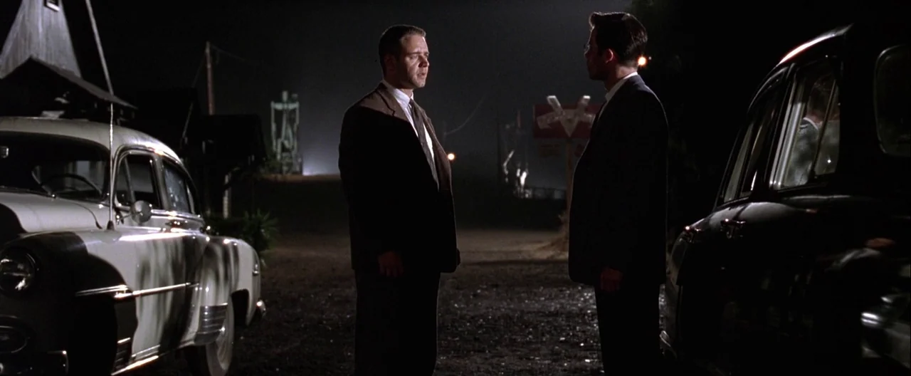 L.A. Confidential (1997) movie still: fullbody — An asphalt ground area at night flanked by two parked vintage 1950s automobiles. Two men in formal…