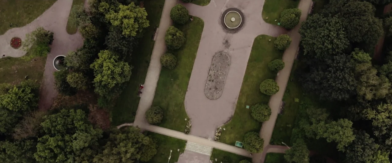 Reagan (2024) movie still: establishing — A public park features walking paths, circular fountains, and dense surrounding trees. The landscape…