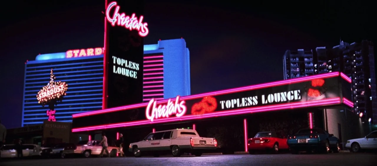 Showgirls (1995) movie still: wide — A nighttime parking lot features a building facade with prominent pink neon signage. Multiple vehicles are…