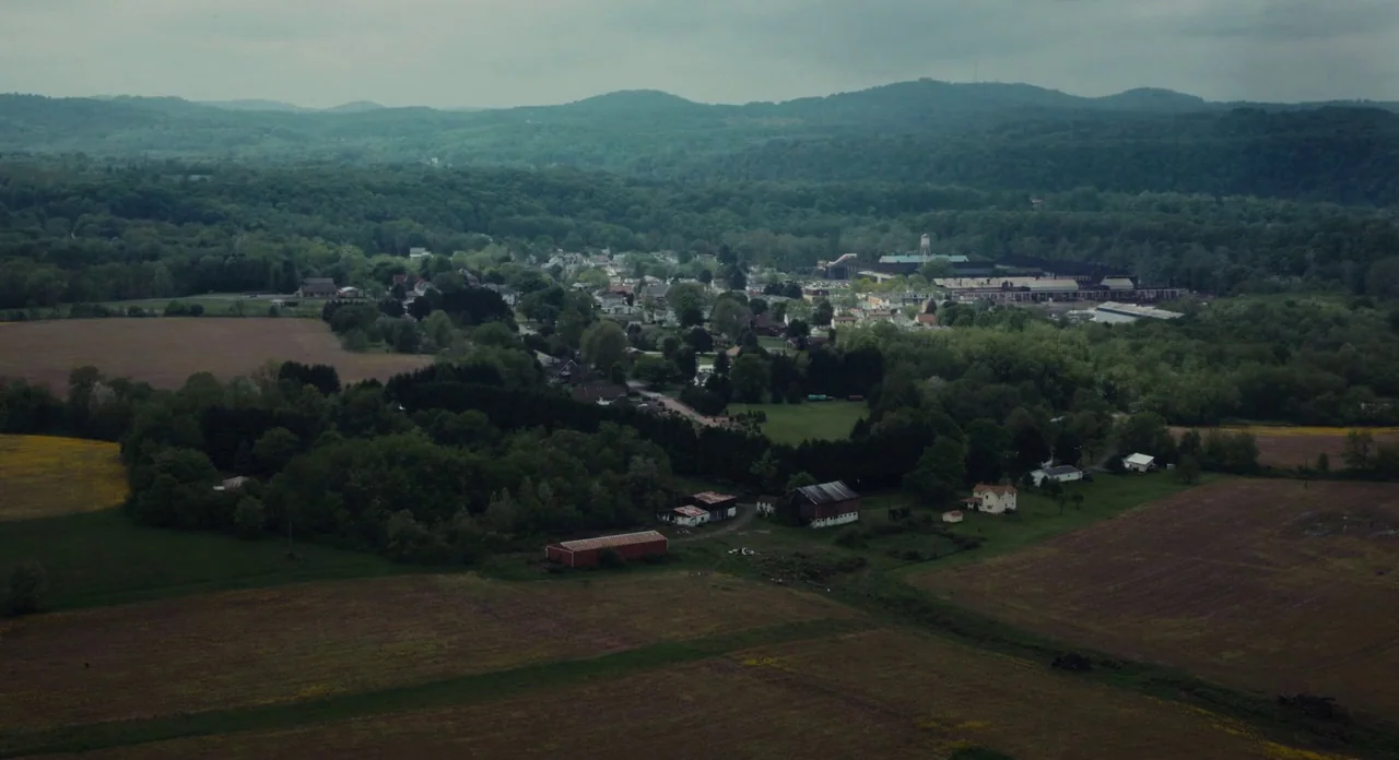 Promised Land (2012) movie still: establishing — A wide aerial view shows a small town nestled within a vast valley. Rolling green hills and…