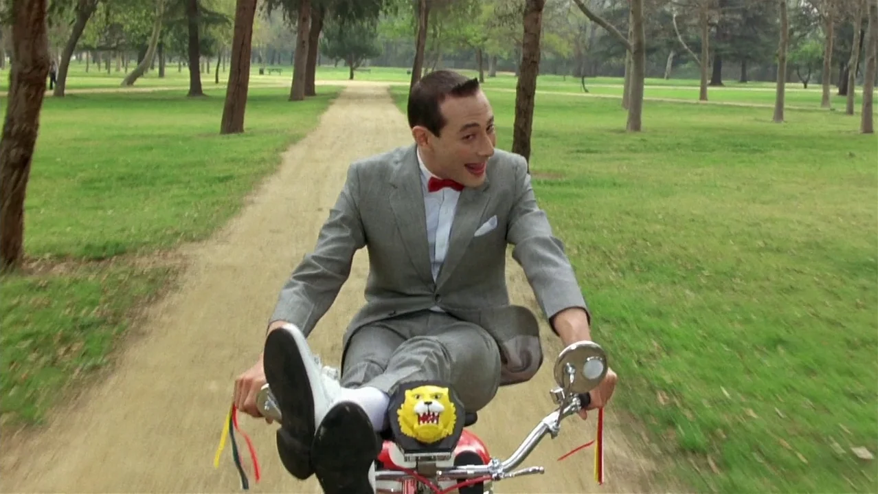 Pee-Wee’s Big Adventure (1985) movie still: fullbody — A man in a grey suit rides a small bicycle along a dirt park path. Rows of trees line the…