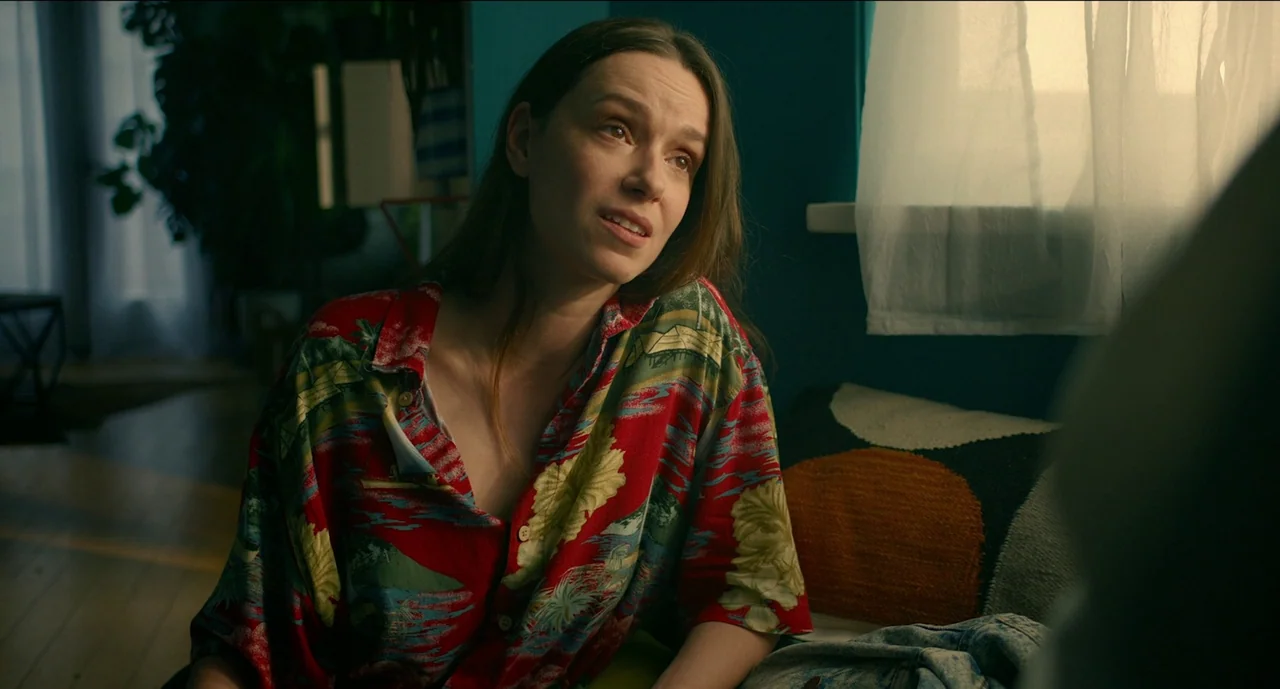 Trim Season (2023) movie still: medium — A woman sits on a sofa against a deep teal wall with a sheer curtained window. She wears a vibrant red…