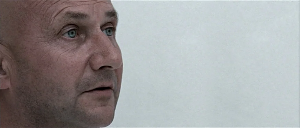 THX 1138 (1971) movie still: closeup — A man's face is positioned on the left against a blank, light-grey wall. He is looking towards the right side…
