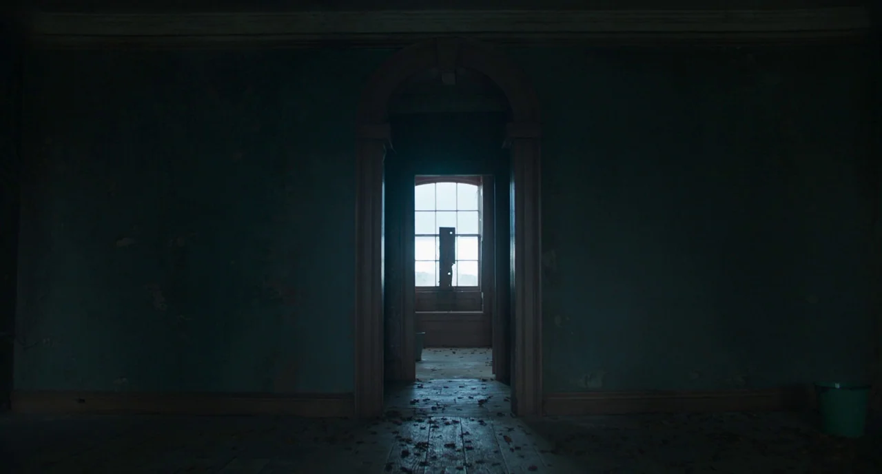The Little Stranger (2018) movie still: establishing — A dark, empty room features a central arched doorway leading to a brighter space. Sunlight…