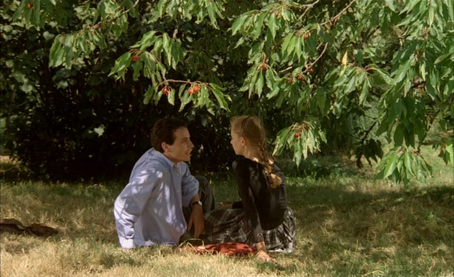 Milou en Mai (1990) movie still: fullbody — A man and a woman sit on the grass beneath a fruit-laden cherry tree. They are facing each other in…