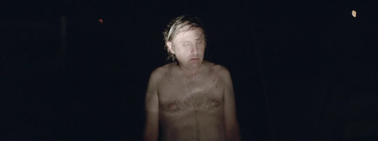 Entertainment (2015) movie still: medium — A man stands centered in complete darkness, his torso illuminated by a focused light. The environment is…