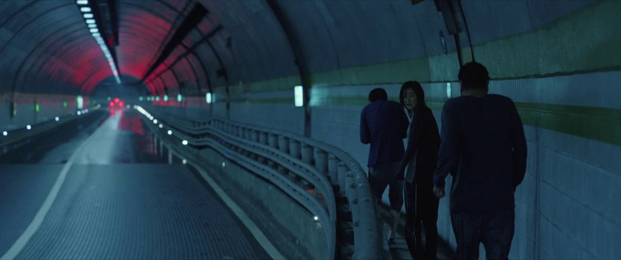 Parasite (2019) movie still: wide — A long, curved concrete road tunnel lit by dim blue lights and distant red signals. Three figures walk along the…